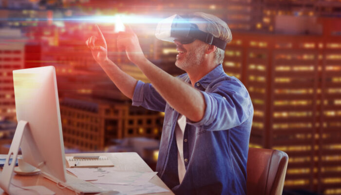 Businessman looking though virtual reality simulator while siting at desk against illuminated buildings in city